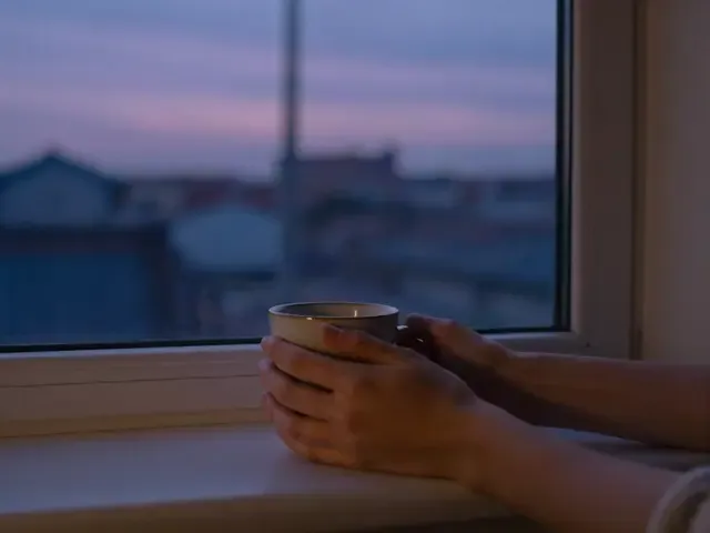 Deux mains tiennent une tasse près d’une fenêtre, à la tombée du jour.